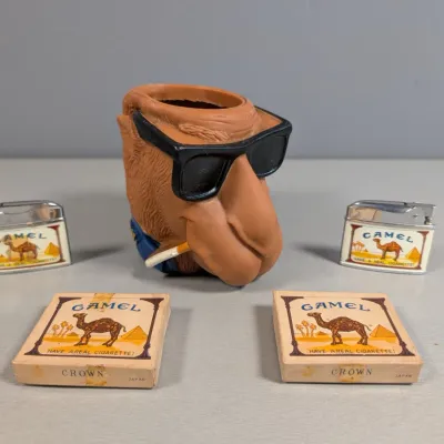 Sale Photo Thumbnail #346: Vintage Joe Camel collectible set featuring a vinyl camel head lighter holder, two flip-top pocket lighters, and two matching Crown Japan matchboxes.