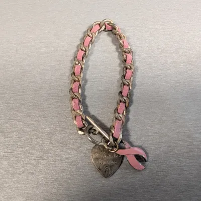Sale Photo Thumbnail #436: Breast cancer awareness charm bracelet featuring a silver-tone chain woven with pink suede, a toggle clasp, an engraved heart charm, and a pink ribbon charm.