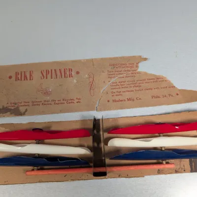 Sale Photo Thumbnail #442: Vintage Bike Spinner novelty toy by Modern Mfg. Co., featuring red, white, and blue plastic propellers mounted on original cardboard packaging.