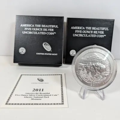 Sale Photo Thumbnail #4: 2011 America the Beautiful Glacier National Park 5 oz silver uncirculated coin, displayed on a small plastic stand with its original United States Mint packaging and Certificate of Authenticity.