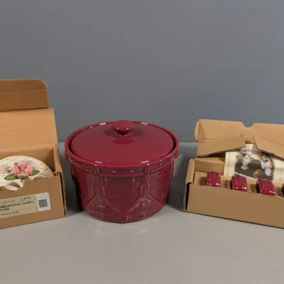 Sale Photo Thumbnail #128: Longaberger pottery items including a maroon basket-weave glazed covered crock, a set of four matching napkin rings in original box, and a flower frog insert with pink dogwood accent in box.