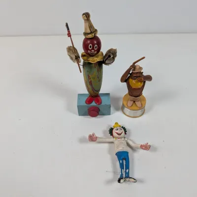 Sale Photo Thumbnail #408: Trio of vintage novelty figures, including a painted wooden clown push-toy, a monkey figurine on a base, and a small plastic clown figure.