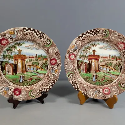Sale Photo Thumbnail #325: Pair of vintage transferware display plates featuring a detailed scenic landscape central motif with brown scrolling floral borders, presented on small display stands.