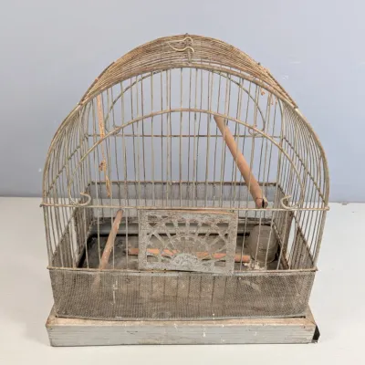 Sale Photo Thumbnail #350: Vintage dome-topped metal birdcage featuring a mesh lower skirt, wooden perches, and decorative front door scrollwork, showing aged patina.