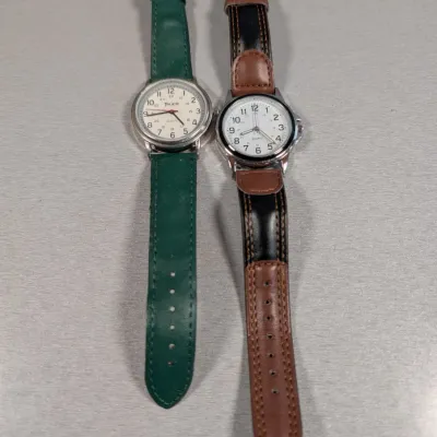 Sale Photo Thumbnail #145: Pair of quartz wristwatches with easy-to-read white analog dials, one featuring a green leather strap and the other a two-tone brown and black leather strap.