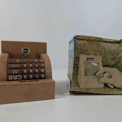 Sale Photo Thumbnail #417: Vintage plastic wind-up cash register toy bank shown alongside its original worn cardboard packaging.