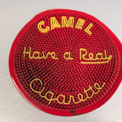 Sale Photo Thumbnail #320: Vintage round red rubber Camel change mat with raised yellow lettering reading "Have a Real Cigarette," showing light wear and surface residue.