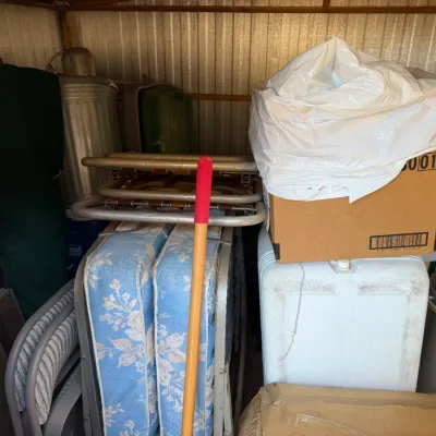 Assorted shed storage items including a foldable cot with floral mattresses, folding webbed lawn chairs, metal trash cans, and cardboard...
