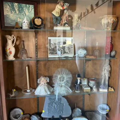 Wooden display cabinet filled with collectibles, including ceramic figurines, geodes, fossils, framed photos, and decorative glassware.