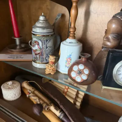 Collection of decorative vintage trinkets and collectibles displayed on glass cabinet shelves, including a lidded beer stein, porcelain...