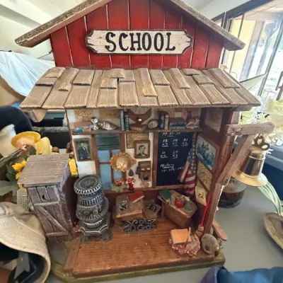 Detailed miniature one-room schoolhouse diorama featuring intricate interior furnishings, a potbelly stove, desks, blackboard, and...