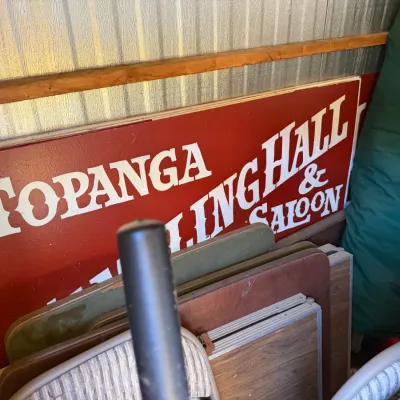 Large vintage painted wooden sign reading 'Topanga Bowling Hall & Saloon', stored alongside folding tables and chairs in a...