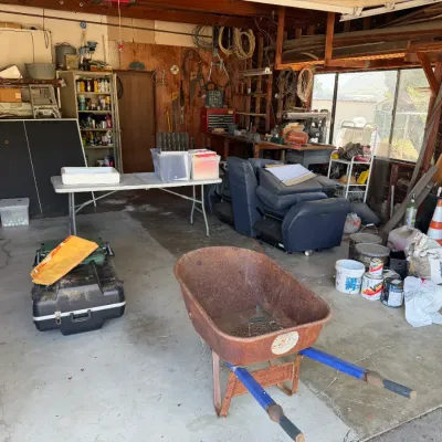 Workshop and garage space filled with tools, a rusted wheelbarrow, folding table, ping pong table, blue recliner, and various home...