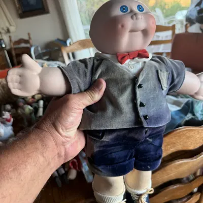 Vintage Cabbage Patch Kids-style porcelain doll wearing a grey vest, red bowtie, navy shorts, and blue shoes.