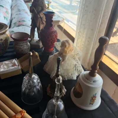 Collection of decorative collectibles including glass and ceramic bells, a carved wooden figurine of a man, a blonde doll in a lace...