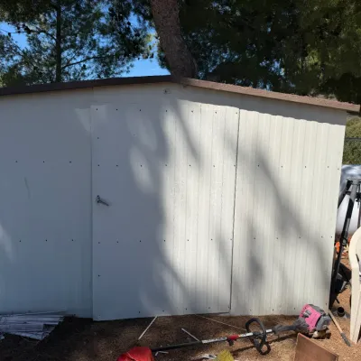 Large white metal outdoor storage shed with double doors and a brown trim roofline, surrounded by outdoor yard tools.