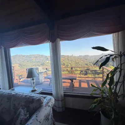 Living room interior with large picture windows overlooking a wooden deck and scenic mountain view, featuring a floral upholstered sofa...
