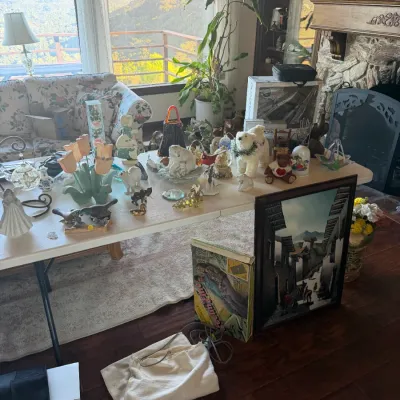 Folding table displaying various collectibles, figurines, glass decor, and framed artwork in a living room with a scenic mountain view.