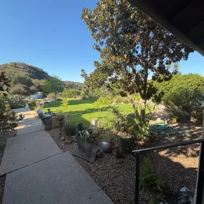Outdoor yard space featuring a concrete walkway, potted plants, trees, and patio seating in a scenic garden setting.