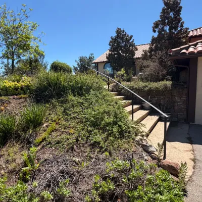 Exterior view of a Spanish-style home featuring a terracotta tile roof, landscaped yard, and stone stairs with a black metal handrail.