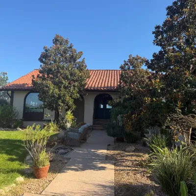 Exterior view of a Spanish-style home featuring a terracotta tile roof, arched entryway, and landscaped front yard with mature trees.