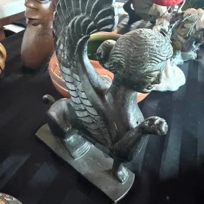 Cast bronze sphinx statue with detailed wings and stylized classical features, displayed alongside other decorative art objects.