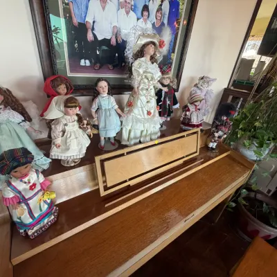 Collection of porcelain and costume dolls in various traditional and character outfits, displayed atop a wood spinet piano.