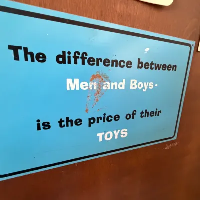Novelty blue metal sign reading 'The difference between Men and Boys- is the price of their TOYS' mounted on a door.