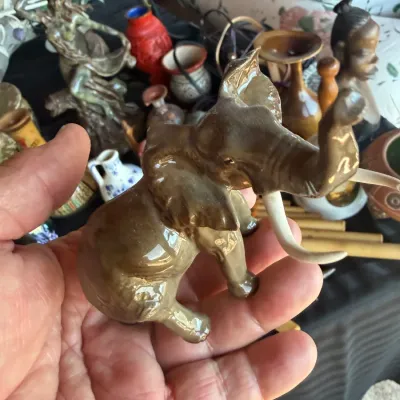 Small glossy brown ceramic elephant figurine with raised trunk and white tusks, held in hand among other collectibles.