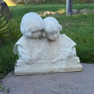 Cast stone garden statue depicting two young children, resting on a flat concrete base with light outdoor weathering.