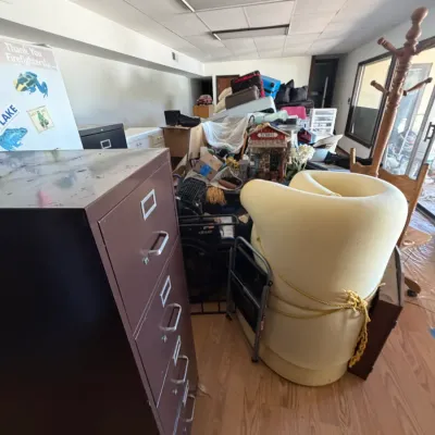 Brown metal four-drawer filing cabinet standing in a crowded room alongside foam seating, a coat rack, and assorted household items.