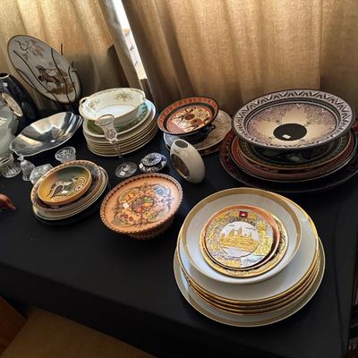 Sale Photo Thumbnail #171: A tabletop display featuring a diverse assortment of decorative plates, bowls, glassware, and souvenir dishes. Items include a gold-rimmed souvenir plate of Cambodia, Greek-style pattern bowls, fine porcelain china, and clear crystal pieces.