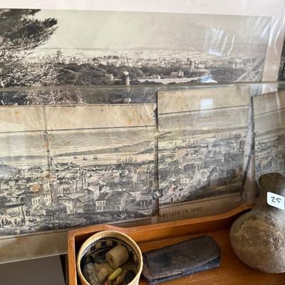 Sale Photo Thumbnail #109: Collection of vintage historical prints, including a framed black-and-white panorama of San Francisco in 1860, displayed alongside small collectibles like a woven basket and a leather case.