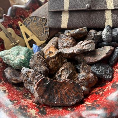 Sale Photo Thumbnail #141: Collection of raw rock specimens and mineral rough displayed in a red-glazed ceramic bowl, alongside a leather glasses case and brass novelties.