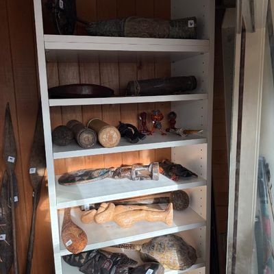 Sale Photo Thumbnail #226: Collection of carved wooden tribal artifacts and figures displayed on white shelving, including masks, clubs, and statues.