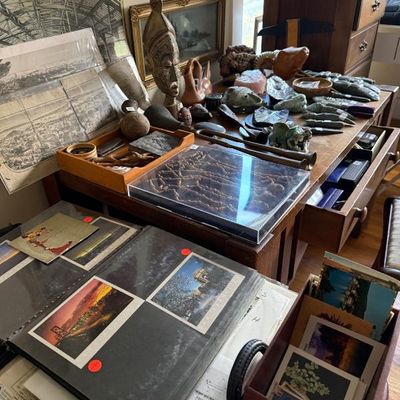 Sale Photo Thumbnail #187: Collection of eclectic collectibles, including vintage photo albums, framed artwork, carved African masks, brass horns, and mineral specimens on a wooden desk.