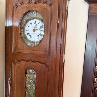 Sale Photo Thumbnail #357: French Provincial style carved oak tall case clock featuring a porcelain dial with Roman numerals and ornate brass accents.