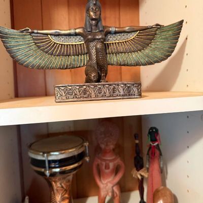 Sale Photo Thumbnail #202: Egyptian-style bronze-finish statue of Isis with outstretched wings, displayed on a shelf above various global figures and a small hand drum.