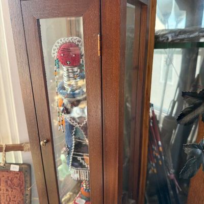 Sale Photo Thumbnail #249: A tall, narrow wooden display cabinet with glass-front door housing a handcrafted beaded doll inside.