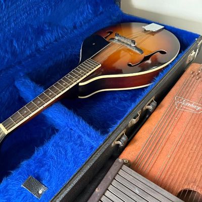Sale Photo Thumbnail #179: A-style sunburst mandolin resting in a blue plush-lined hard shell case alongside an Autoharp.