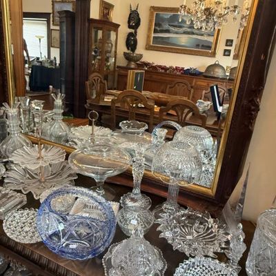 Sale Photo Thumbnail #338: Collection of vintage cut glass and crystal tableware, including a blue cut glass basket, clear candy dishes, decanters, and covered serving pieces, arranged on a dark wood credenza in front of a large mirror.