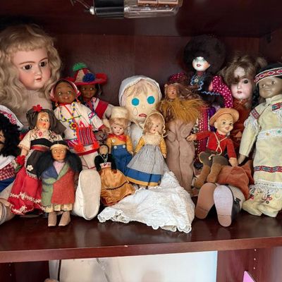 Sale Photo Thumbnail #242: Collection of vintage and antique dolls, including bisque-head, rag, and multicultural souvenir dolls, displayed on a wooden shelf.