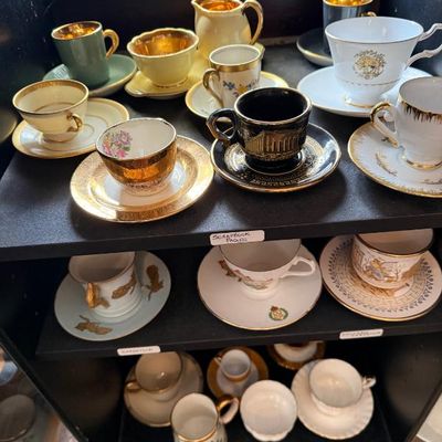 Sale Photo Thumbnail #328: Collection of vintage porcelain teacups, saucers, and creamers displayed on dark shelving, featuring gold-gilt trim and ornate painted details.