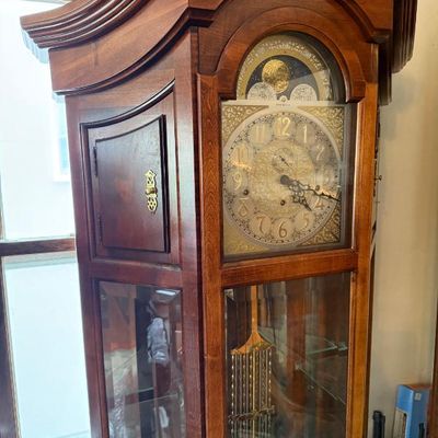 Sale Photo Thumbnail #358: Traditional wooden grandfather clock with an ornate brass dial, moon phase display, and glass display shelving inside the base.