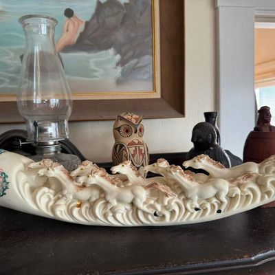 Sale Photo Thumbnail #263: Faux-ivory tusk carving depicting galloping horses, displayed alongside a clear glass oil lamp, a painted ceramic owl figurine, and various decorative items.