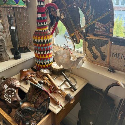 Sale Photo Thumbnail #229: Collection of global ethnographic decor, featuring a tall colorful beadwork jug, carved wooden tribal masks, hand percussion instruments, and a large woven horse head figure.