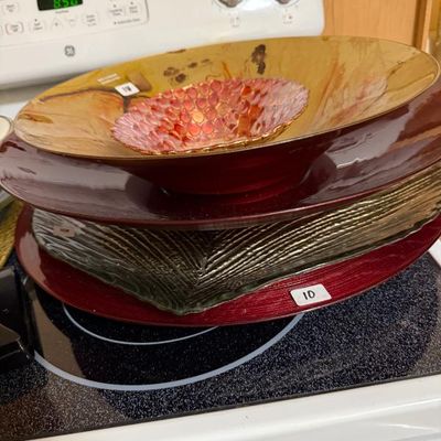 Sale Photo Thumbnail #168: Stack of decorative serving bowls and trays, including a red textured charger plate, a textured glass platter, and a gold-toned painted bowl.