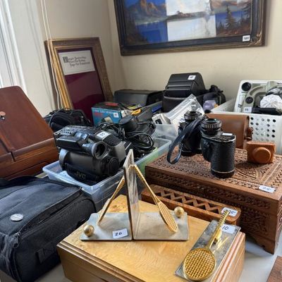 Sale Photo Thumbnail #188: Collection of vintage items featuring brass tennis racquet bookends, a JVC camcorder, carved wooden boxes, and binoculars displayed on a table.