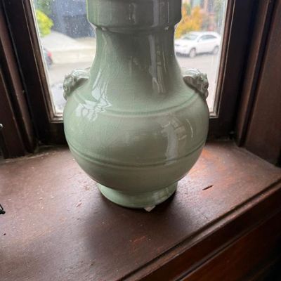 Sale Photo Thumbnail #297: Celadon green ceramic vase featuring a crackle glaze and molded lion-head handles, set on a wooden windowsill.