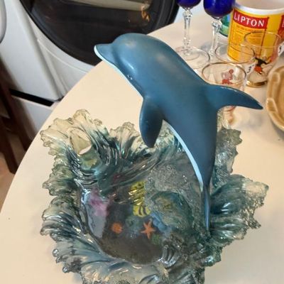 Sale Photo Thumbnail #310: Leaping dolphin figurine set on a clear blue resin wave base with an underwater reef scene, shown alongside cobalt blue stemware and vintage drinking glasses.